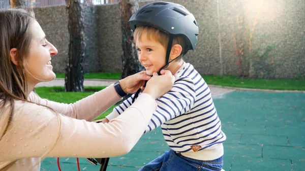 Portret uśmiechniętej młodej matki regulującej i zapiętej klamry na hełm ochronny syna przed jazdą na rowerze — Zdjęcie stockowe