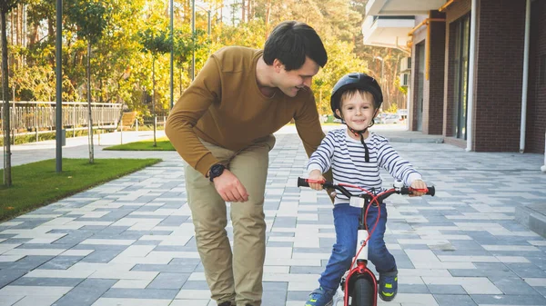 Image tonique de heureux petit garçon souriant chevauchant son premier vélo avec son père marchant près de lui — Photo