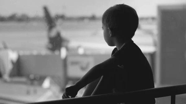 Black and white upset little boy sitting in airport terminal and looking on big airplanes — Stock Photo, Image