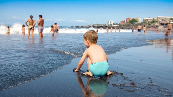 Mały chłopczyk siedzi na plaży i bawi się falami oceanu. — Zdjęcie stockowe