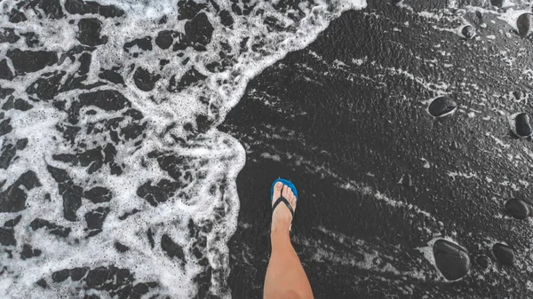黒い火山砂の海のビーチを歩くフリップフロップでオスの足の上からの眺め — ストック写真