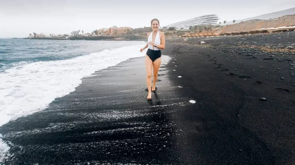 Beautfiul smiling young woman in swimsuit running on the sea beach with black volcanic sand — Stock Photo, Image