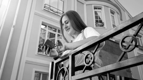 Portrait of beautiful smiling woman standing on balcony and typing message on smartphone — Stock Photo, Image