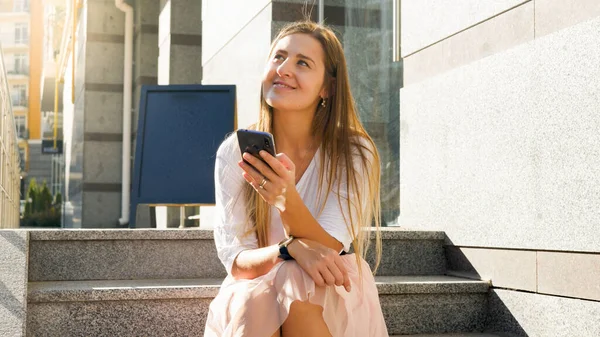Портрет улыбающейся мечтательной молодой женщины, сидящей на каменной лестнице на городской улице и держащей мобильный телефон — стоковое фото