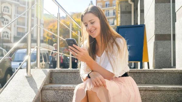 Toned fénykép boldog mosolygós lány ül a lépcsőn az utcán, és böngészi az interneten okostelefon — Stock Fotó