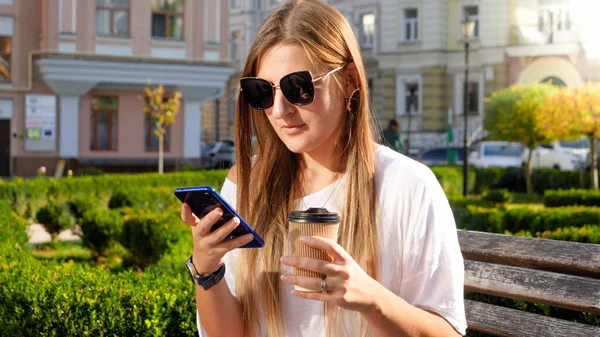 Retrato de una estudiante bebiendo café y usando un smartphone en el parque —  Fotos de Stock