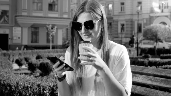 Черно-белый портрет улыбающейся девушки, пьющей кофе и просматривающей интернет на смартфоне, сидящей на скамейке в парке — стоковое фото