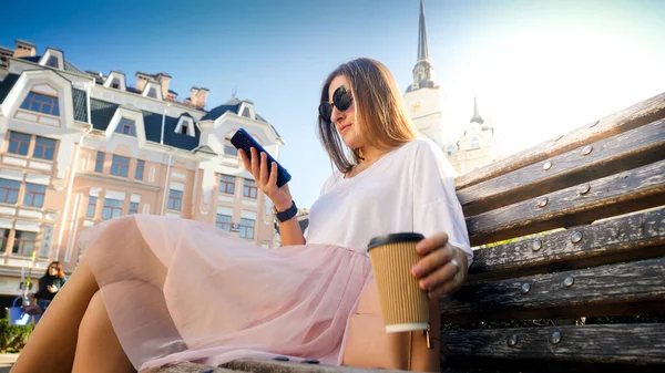Jovem mulher bonita bebendo café e digitando mensagem no smartphone enquanto sentado no banco no jardim — Fotografia de Stock