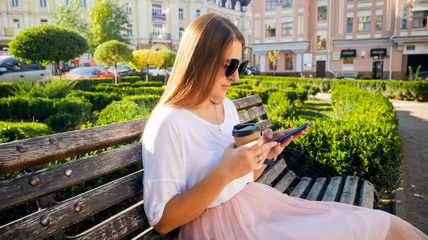 Beautiful young woman in park using smartphone and drinking coffee — Stock Photo, Image