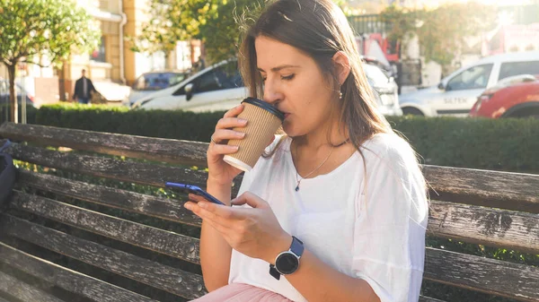 Тонированный портрет молодой студентки, пьющей кофе и просматривающей интернет на смартфоне в парке — стоковое фото