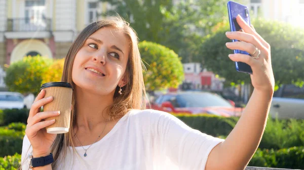 コーヒーカップで笑顔の女性の肖像画は、公園でスマートフォンで自撮り写真を作る — ストック写真