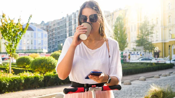 Retrato de hermosa chica sonriente en gafas de sol bebiendo café y montando en scooter eléctrico en la calle de la ciudad —  Fotos de Stock