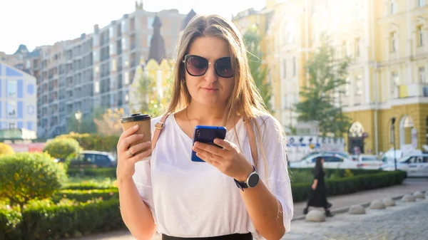 Retrato de bela jovem cabelo comprido segurando xícara de café andando na rua e digitando mensagem nas mídias sociais no smartphone — Fotografia de Stock