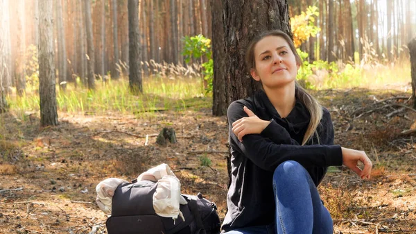 Mutlu gülümseyen genç bir kadının portresi yerde oturuyor ve ormanda çam ağacına yaslanıyor. — Stok fotoğraf