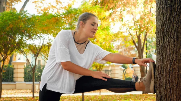Portrait of sporty young woman stretching her muscles before doing sports exercises in autumn park — Stock Photo, Image
