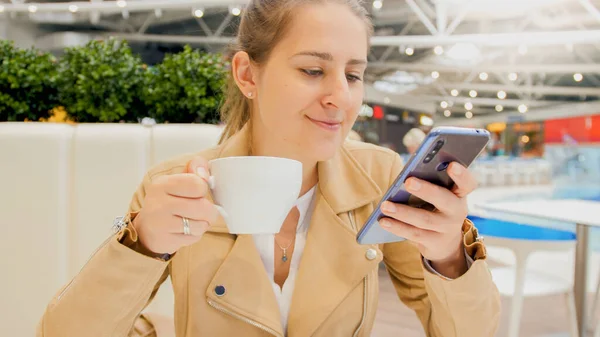 Retrato de jovem sorridente lendo notícias no smartphone enquanto toma café da manhã no café — Fotografia de Stock