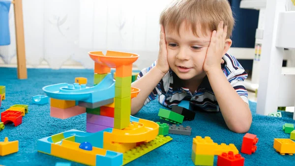 Portrait of cute toddler boy lying on floor and looking on colorful construction from plastic bricks and blocks — Stock Photo, Image