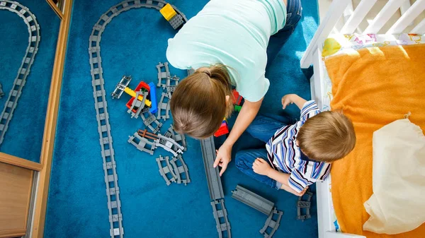 Vista dall'alto su giovane madre con piccolo figlio che costruisce ferrovia giocattolo da mattoni e blocchi colorati — Foto Stock