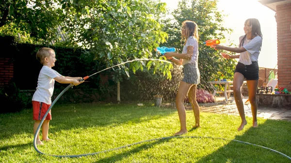 İki kız kardeşi olan küçük bir çocuk arka bahçede su tabancası ve hortumla oynuyor. — Stok fotoğraf
