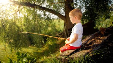 Nehirdeki bankta oturan sevimli küçük çocuk ve eski ahşap çubukla balık tutuyor.