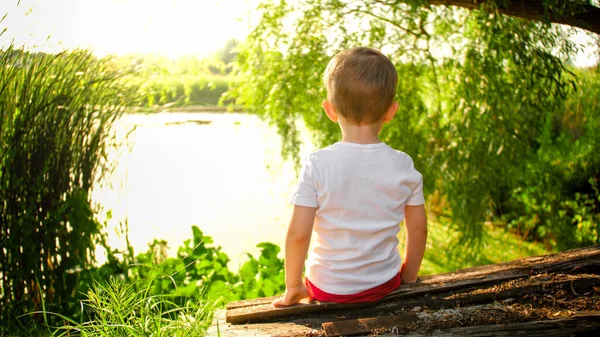 Achteraanzicht phto van kleine jongen zittend op de houten bank bij het meer en kijkend op rustig water — Stockfoto