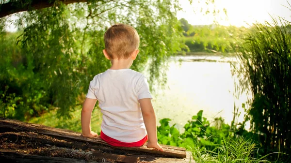 Маленький мальчик сидит в деревне у озера и смотрит на закат солнца — стоковое фото