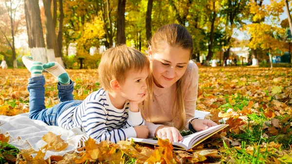 Portrait of little boy lying with his mother at autumn park and reading a book — Stock Photo, Image