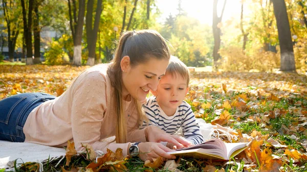 Portrait d'une jeune mère racontant une histoire à son petit fils allongé sur des feuilles d'arbres jaunes tombées au parc d'automne — Photo