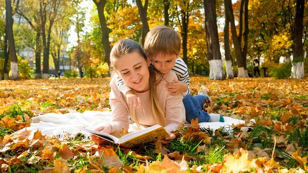 Förtjusande liten pojke sitter på mammor tillbaka och läser en bok i höstparken — Stockfoto