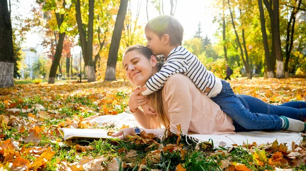 在秋天的公园里，一个蹒跚学步的小男孩坐在妈妈的背上亲吻着她 — 图库照片