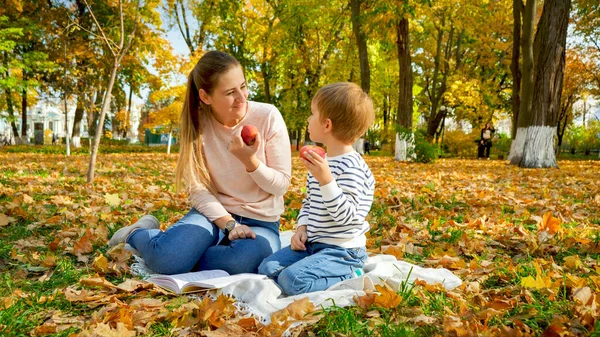 Retrato de una joven madre sonriente con su pequeño hijo haciendo un picnic en una manta en el parque de otoño — Foto de Stock