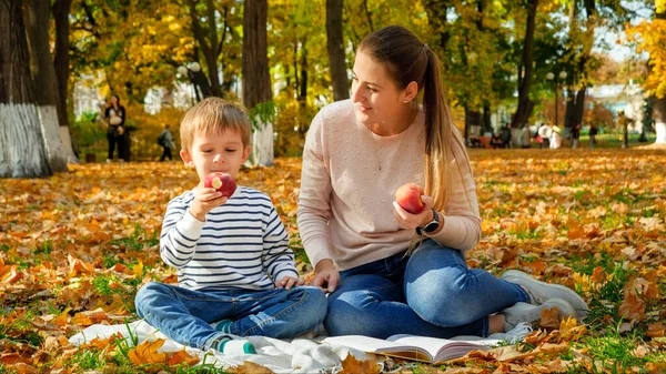 Kleiner Junge mit schöner junger Mutter sitzt auf dem Boden im Herbstpark und isst roten Apfel — Stockfoto
