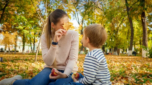 Schöne junge Frau blickt durch gelbes Herbstblatt auf ihren Sohn im Herbstpark — Stockfoto