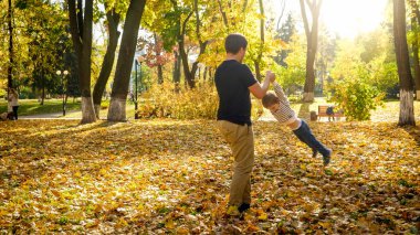 Genç baba oğlunu elleriyle tutup sonbahar parkında dönüyor.