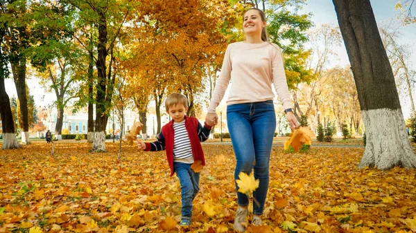 Счастливая улыбающаяся молодая мать держит сына за руку и гуляет по осеннему парку — стоковое фото