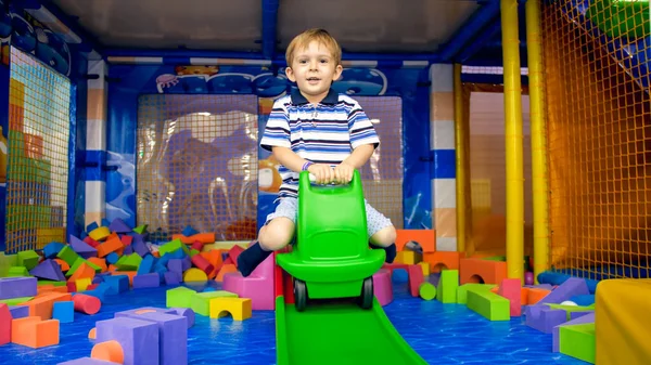 Feliz sorrindo menino cavalgando a corrediça no carrinho de plástico de brinquedo na sala de jogos no parque de diversões — Fotografia de Stock