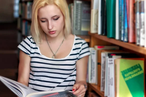 Jong blond meisje met open boek in de hand staat onder de boekenkasten — Stockfoto