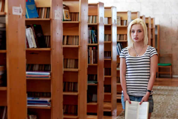 Jong blond meisje staat met open boek in de hand onder de boekenkasten — Stockfoto