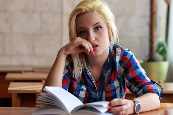 Ontevreden blond meisje in geruite hemd met een open boek zit aan een bureau — Stockfoto