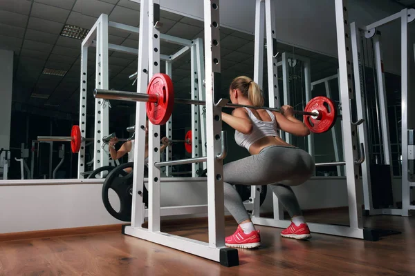 Joven sexy rubia europea chica en el gimnasio haciendo sentadilla con barra en frente del espejo — Foto de Stock