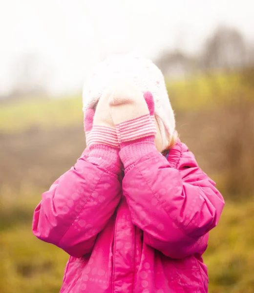 A child in a red jacket and a white hat plays hide and seek on the street. Autumn. The face is covered with mittens. — Stock Photo, Image