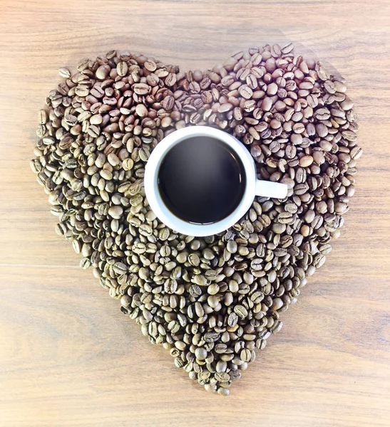 Das Herz der Kaffeebohnen auf einem hölzernen Hintergrund in der Mitte einer weißen Tasse mit Kaffee, mit ihm kommt Dampf. — Stockfoto