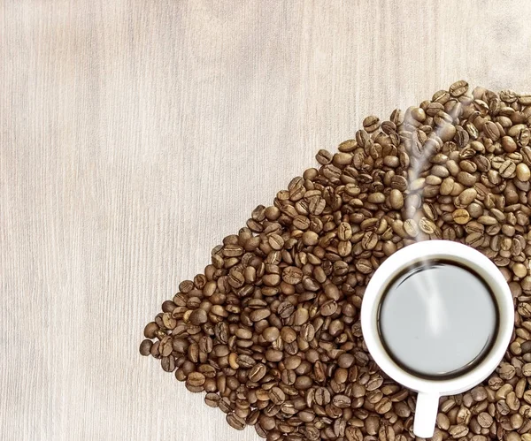 Das Herz der Kaffeebohnen auf einem hölzernen Hintergrund in der Mitte einer weißen Tasse mit Kaffee, mit ihm kommt Dampf. — Stockfoto