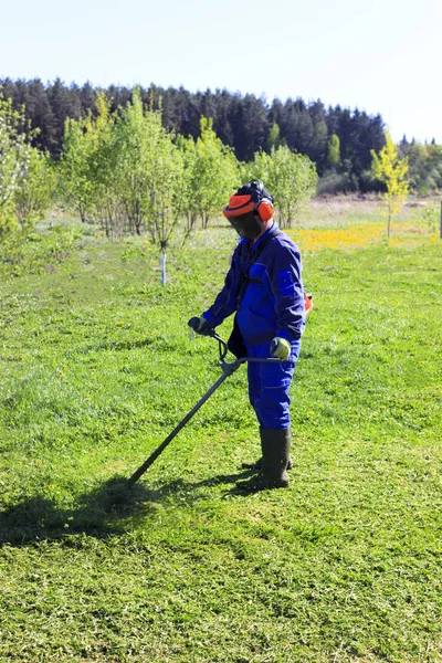 Un hombre con un traje azul. En un casco protector y auriculares. Produce la cosecha de hierbas. La cara no se enfoca — Foto de Stock