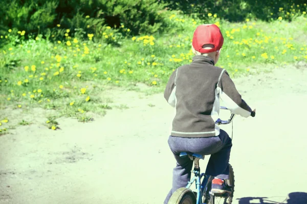 Uma velha estrada rural. Um menino com uma camisola marrom e um chapéu de beisebol vermelho está dirigindo ao longo da estrada em uma bicicleta retro. Visão traseira — Fotografia de Stock