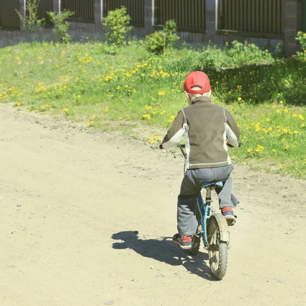 Uma velha estrada rural. Um menino com uma camisola marrom e um chapéu de beisebol vermelho está dirigindo ao longo da estrada em uma bicicleta retro. Visão traseira — Fotografia de Stock