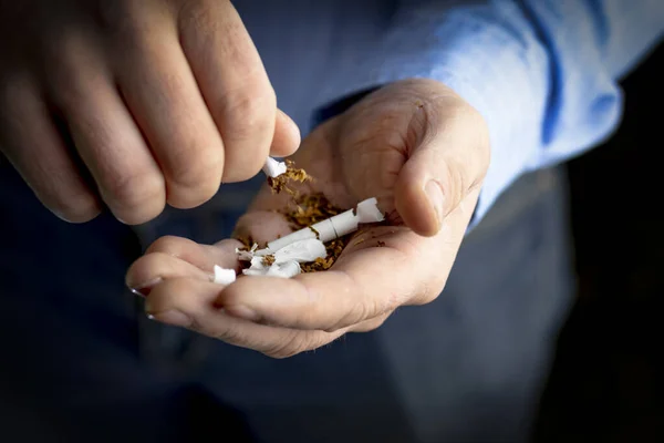 man in a blue shirt, dark background. he has a broken cigarette in his hand. he spills tobacco