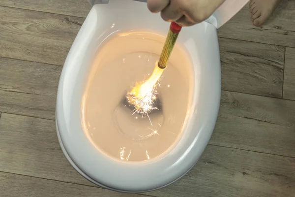 White toilet bowl on a wooden background. the hand lowers the lit firecracker. close-up. — Stock Photo, Image