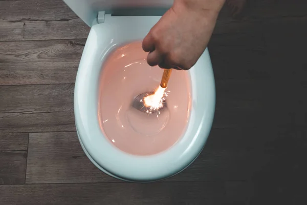 Λευκή λεκάνη τουαλέτας σε ξύλινο φόντο. Το χέρι χαμηλώνει το αναμμένο πυροτέχνημα. κοντινό πλάνο. — Φωτογραφία Αρχείου