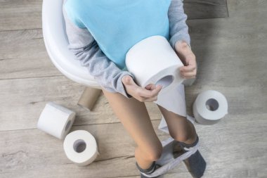 Çocuk beyaz bir tuvalette oturuyor. Elinde tuvalet kağıdı var. Yerde bir sürü rulo var. yakın plan.